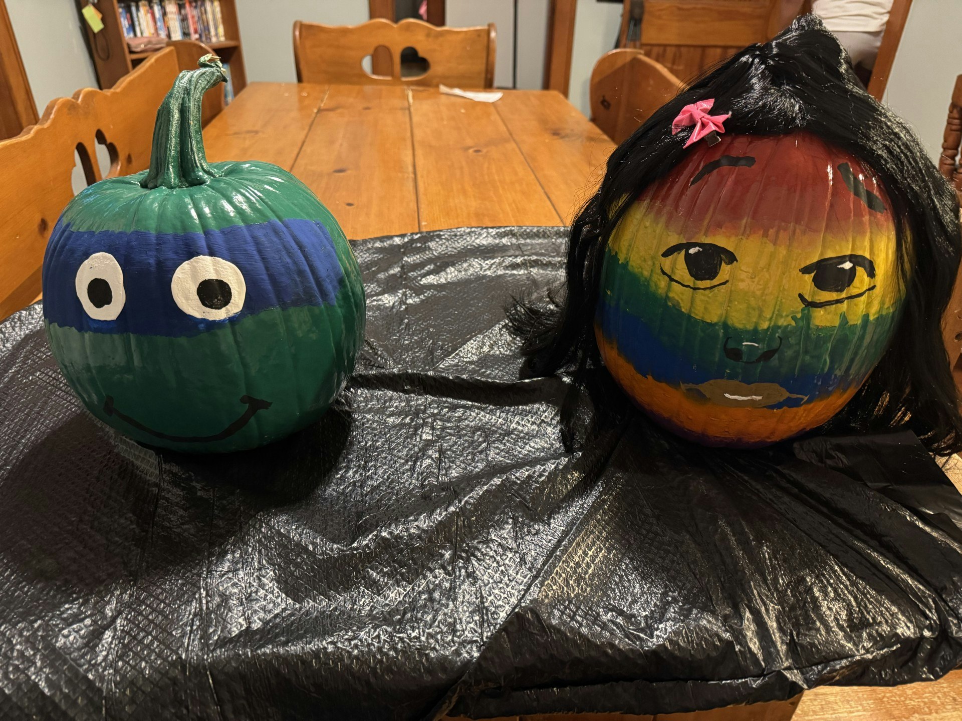 Two painted pumpkins: one as a green turtle with blue mask, the other with a colorful face and wig, on a table.