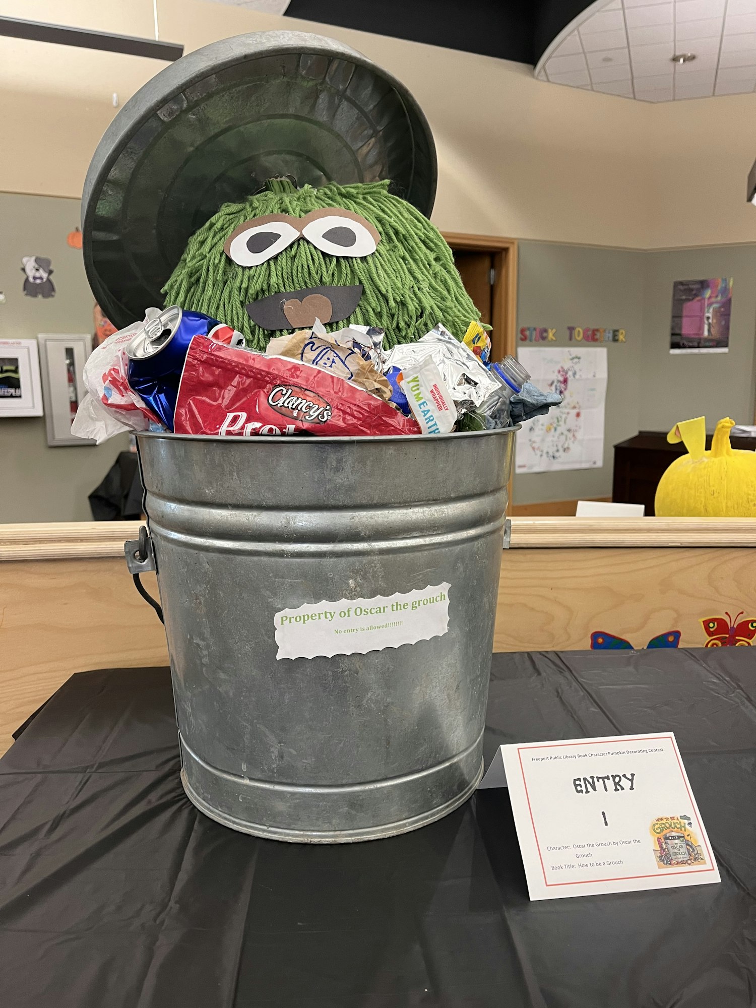 Green puppet resembling a muppet character in a trash can filled with snack wrappers and a label saying "property of Oscar the grouch."