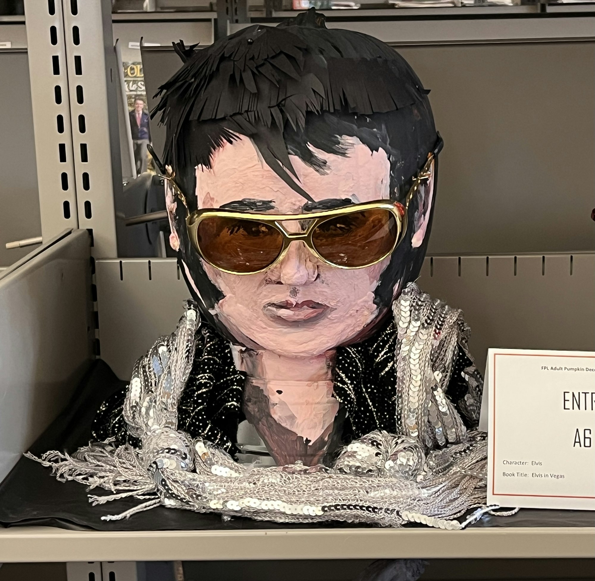A painted pumpkin resembling a person in sunglasses and a sequined outfit, displayed on a shelf with a label beside it.