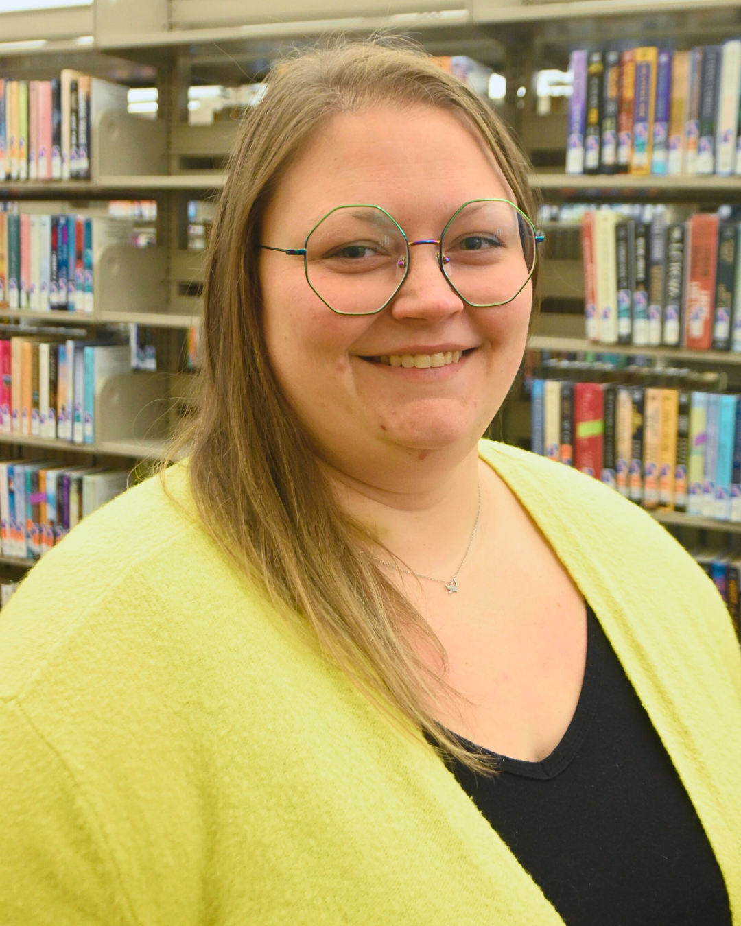 A smiling person with glasses and long hair, wearing a yellow sweater, stands in front of bookshelves in a library.