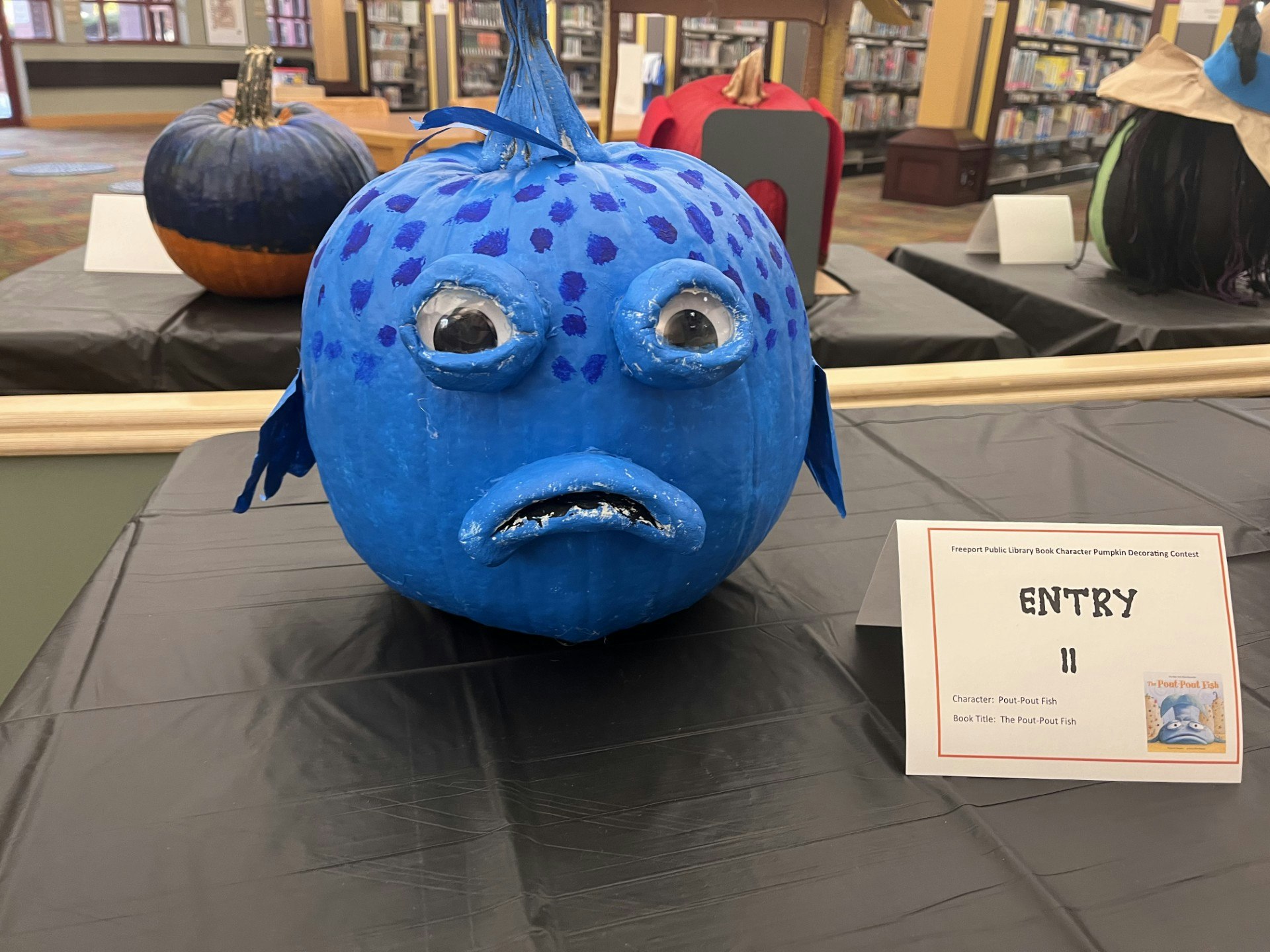 A blue pumpkin decorated as a fish with a sign reading "ENTRY 11," part of a library's book character pumpkin contest.