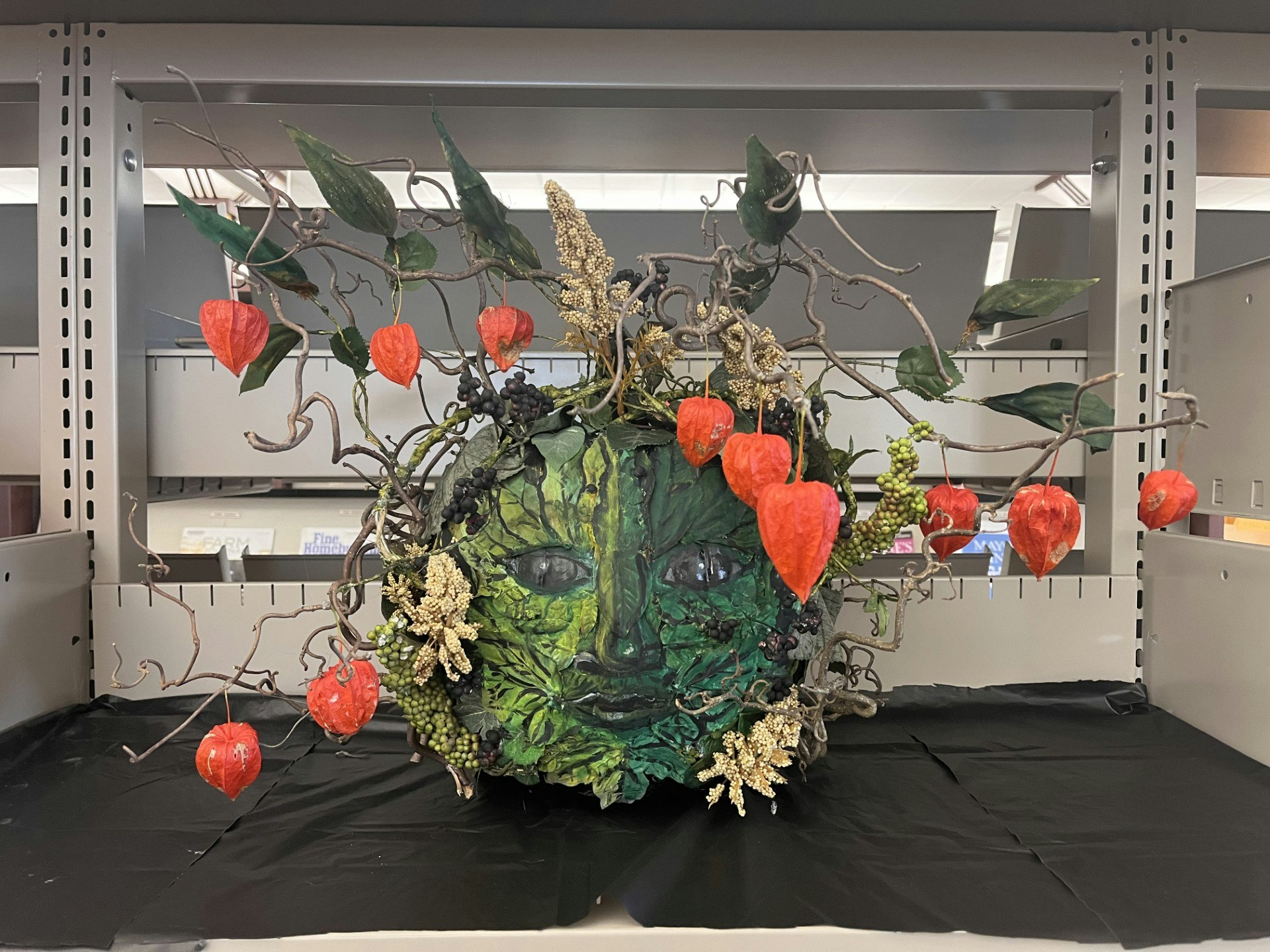 A green face with leaves, branches, and red lantern-shaped ornaments on a black surface.