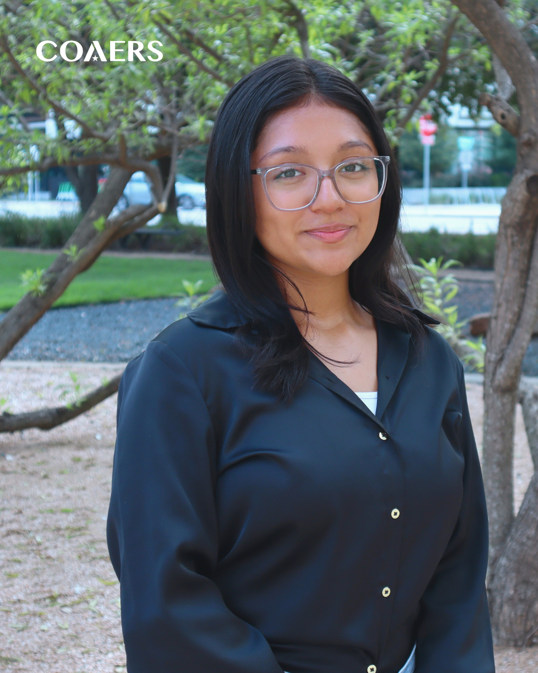 A person with glasses and long hair, wearing a dark shirt, standing outdoors with trees and a path in the background. Text says "COAERS."