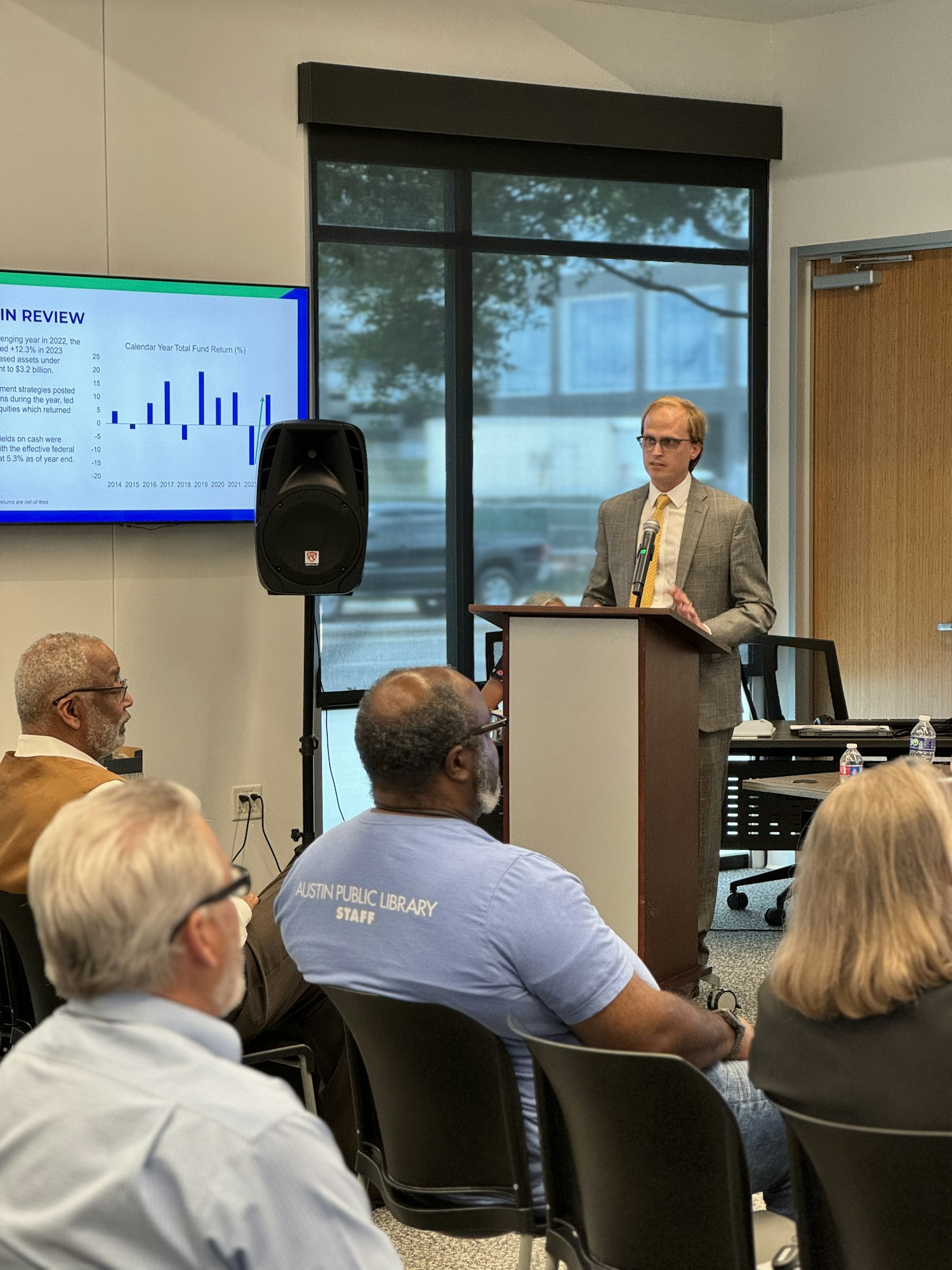 A presentation in progress with an audience and a speaker at a podium; a financial chart is displayed on a screen.
