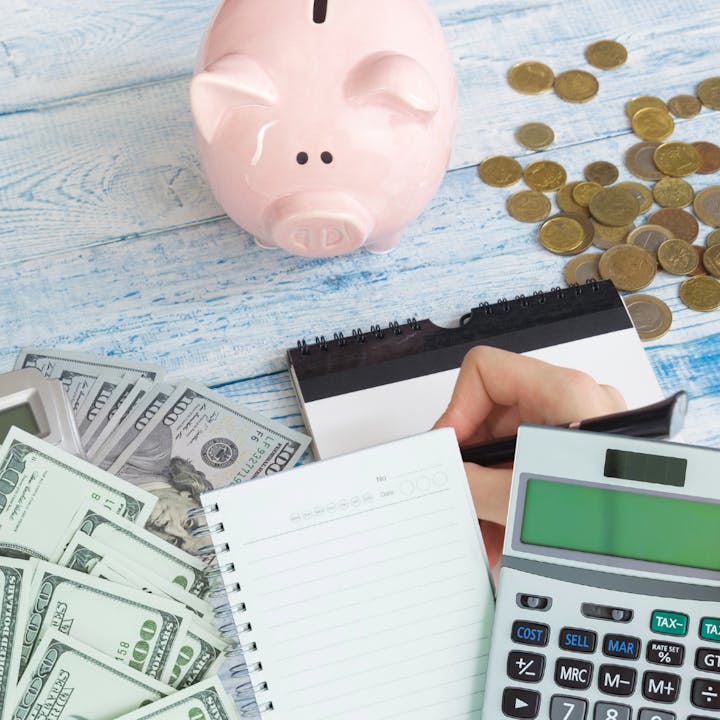 Piggy bank, coins, dollar bills, notebook, calculator, and a hand writing on a notepad on a wooden surface.