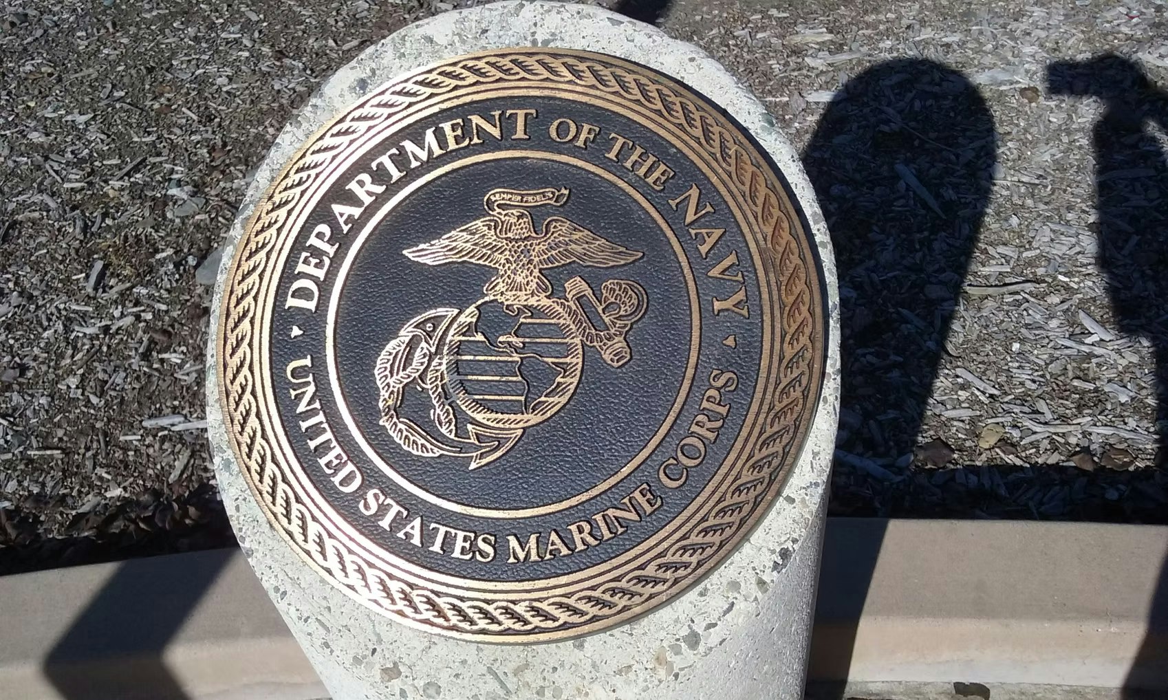 The image shows a bronze emblem of the U.S. Marine Corps, part of the Department of the Navy, mounted on a concrete base.