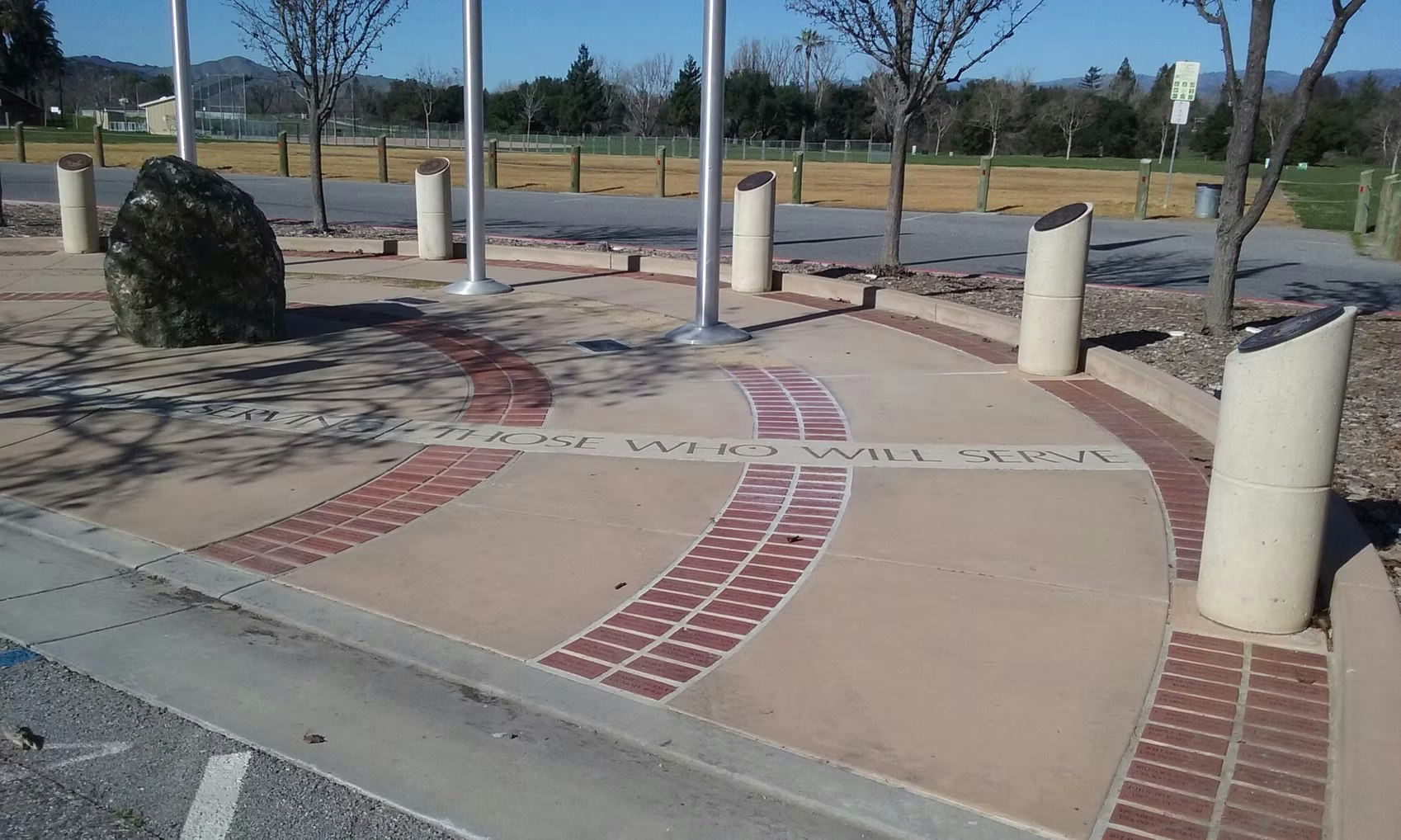 The image shows a plaza with a large rock, flagpoles, and a pathway inscribed with the phrase "DEDICATED TO THOSE WHO WILL SERVE."