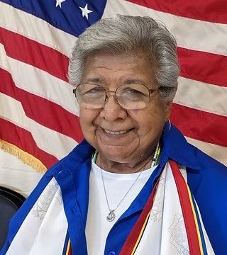 An older woman with gray hair and glasses smiles in front of an American flag, wearing a blue shirt and colorful ribbons.