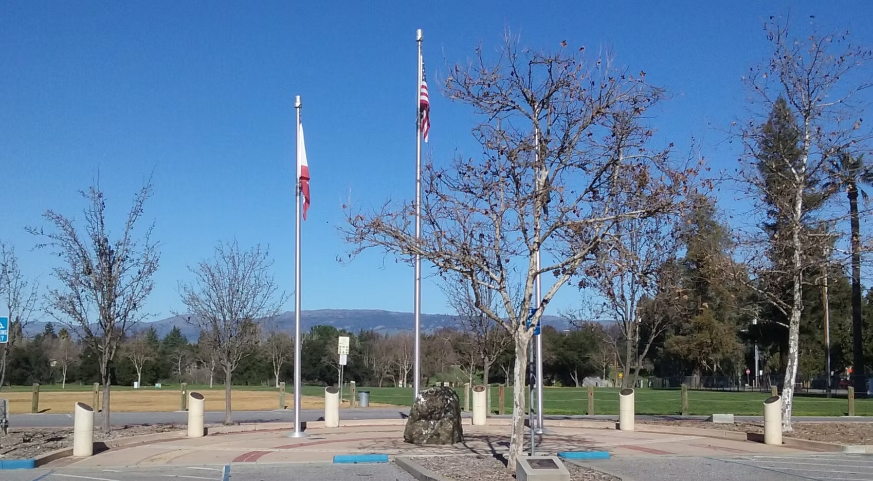 The image shows a park area with bare trees, two flagpoles (one with a California flag), and a distant mountain backdrop under a clear blue sky.
