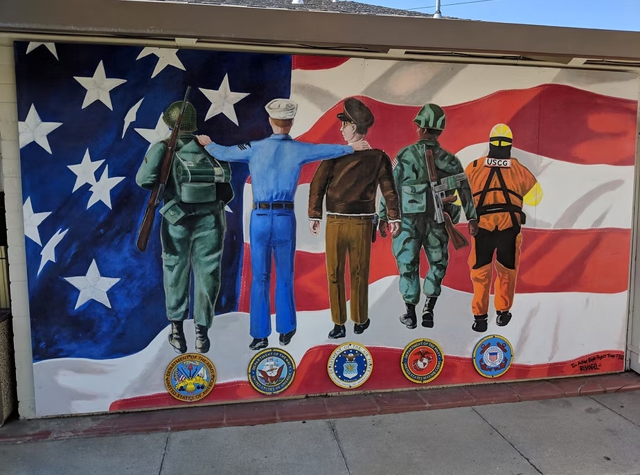 The image shows a mural depicting five military personnel against a backdrop of the American flag, highlighting their service.