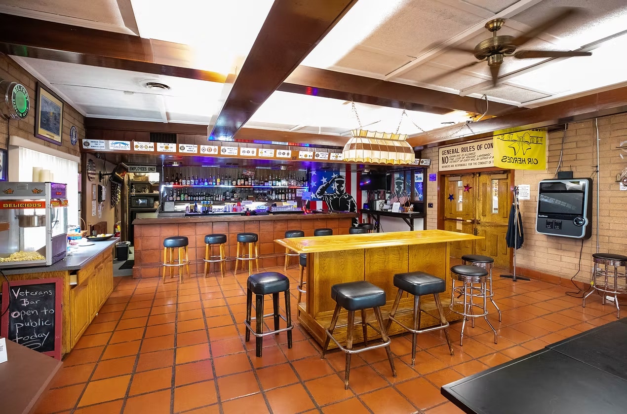A cozy bar interior featuring a wooden bar, stools, a popcorn machine, and a variety of drinks on display.