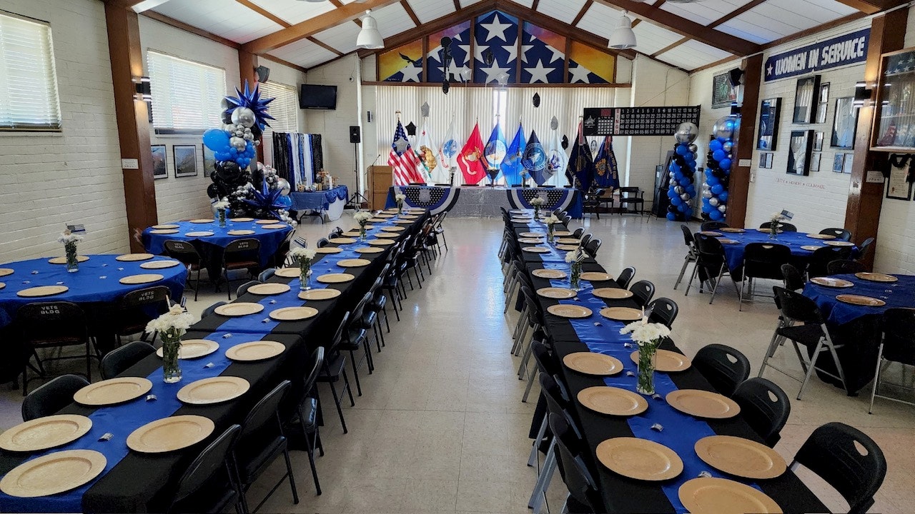 The image shows a nicely arranged banquet hall with blue tablecloths, empty plates, floral centerpieces, and military flags.