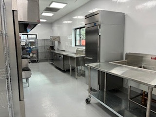 A clean and modern kitchen with stainless steel shelves, counters, and a refrigerator, featuring bright lighting and tiled flooring.