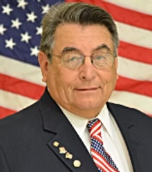 A man in a suit with glasses, standing in front of an American flag, wearing a patriotic tie and medals.