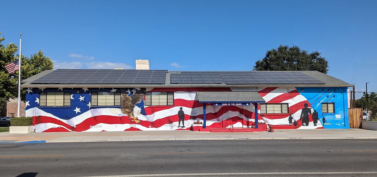 A mural on a building features an American flag design, an eagle, and silhouettes of military figures, along with solar panels.