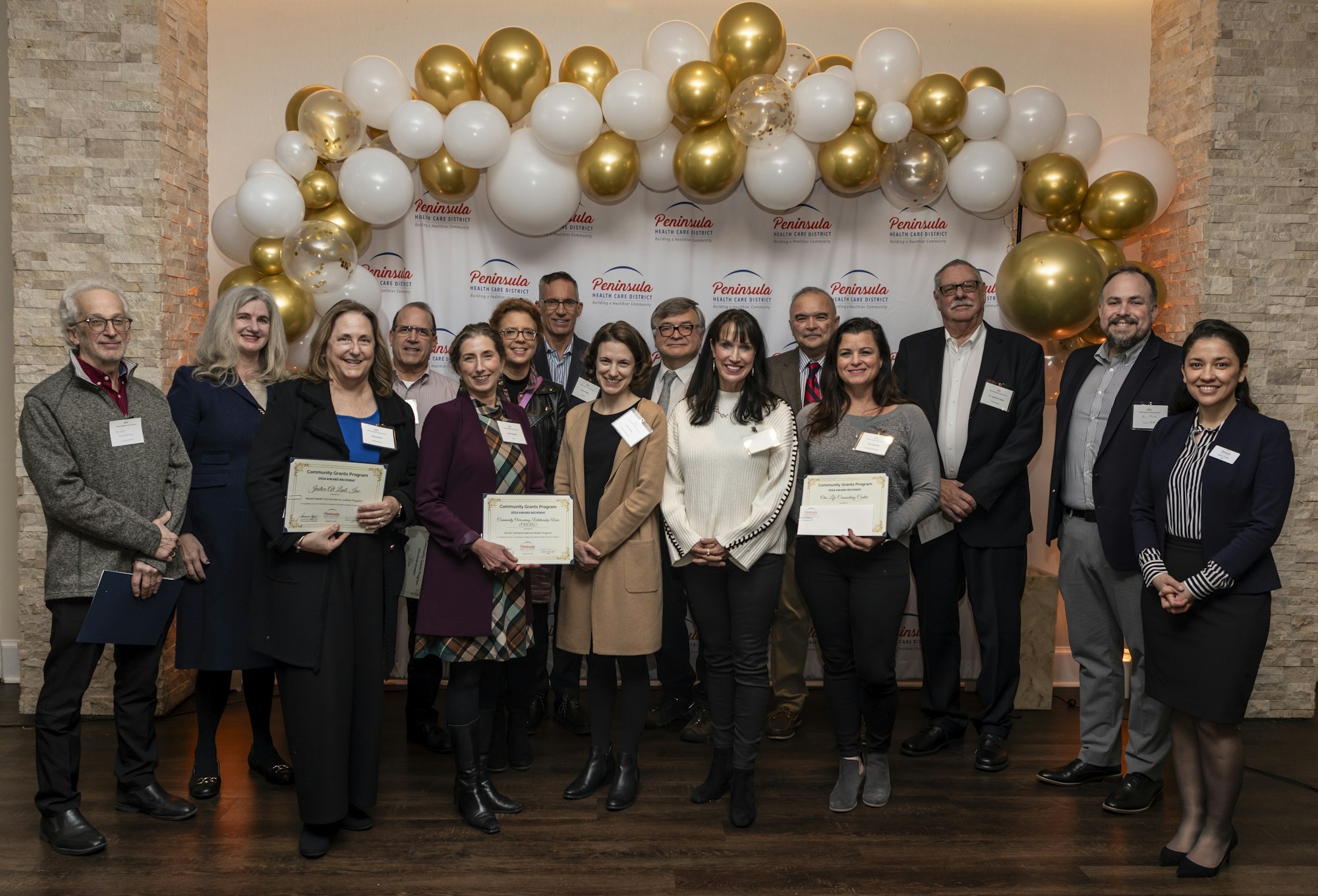 Group of smiling people posing with certificates, balloons in the background, likely a recognition or award event.