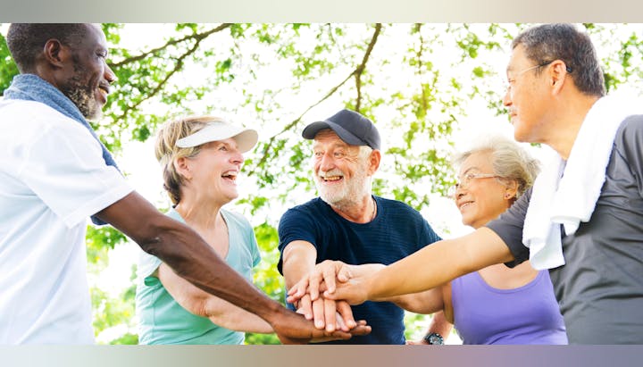 A group of five older adults smiling, stacking hands outdoors in a circle under green trees.