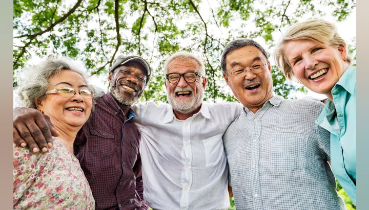 A group of five older adults smiling and embracing, standing outdoors under trees.