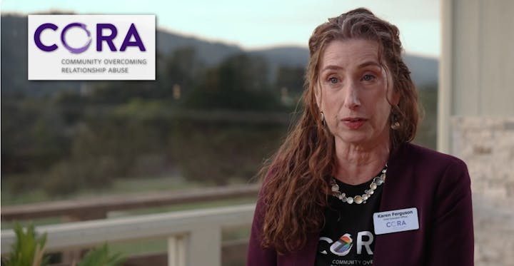 A person with a name tag, wearing a "CORA" shirt, and a "Community Overcoming Relationship Abuse" sign is in the background.