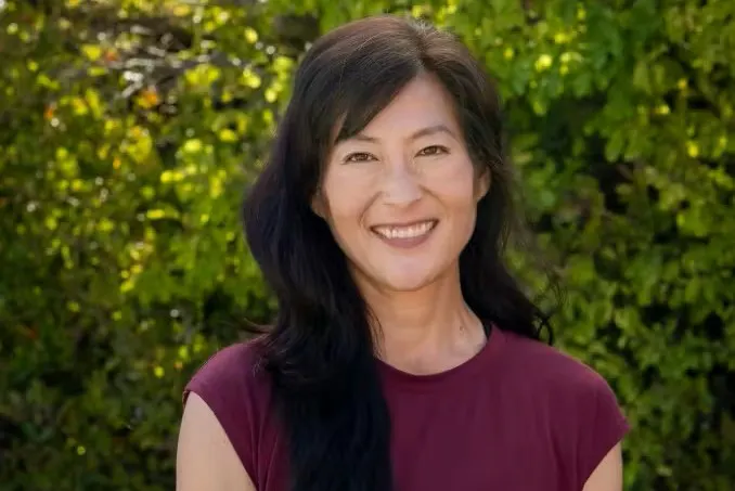 A smiling woman with long dark hair, wearing a maroon top, stands against a backdrop of greenery.