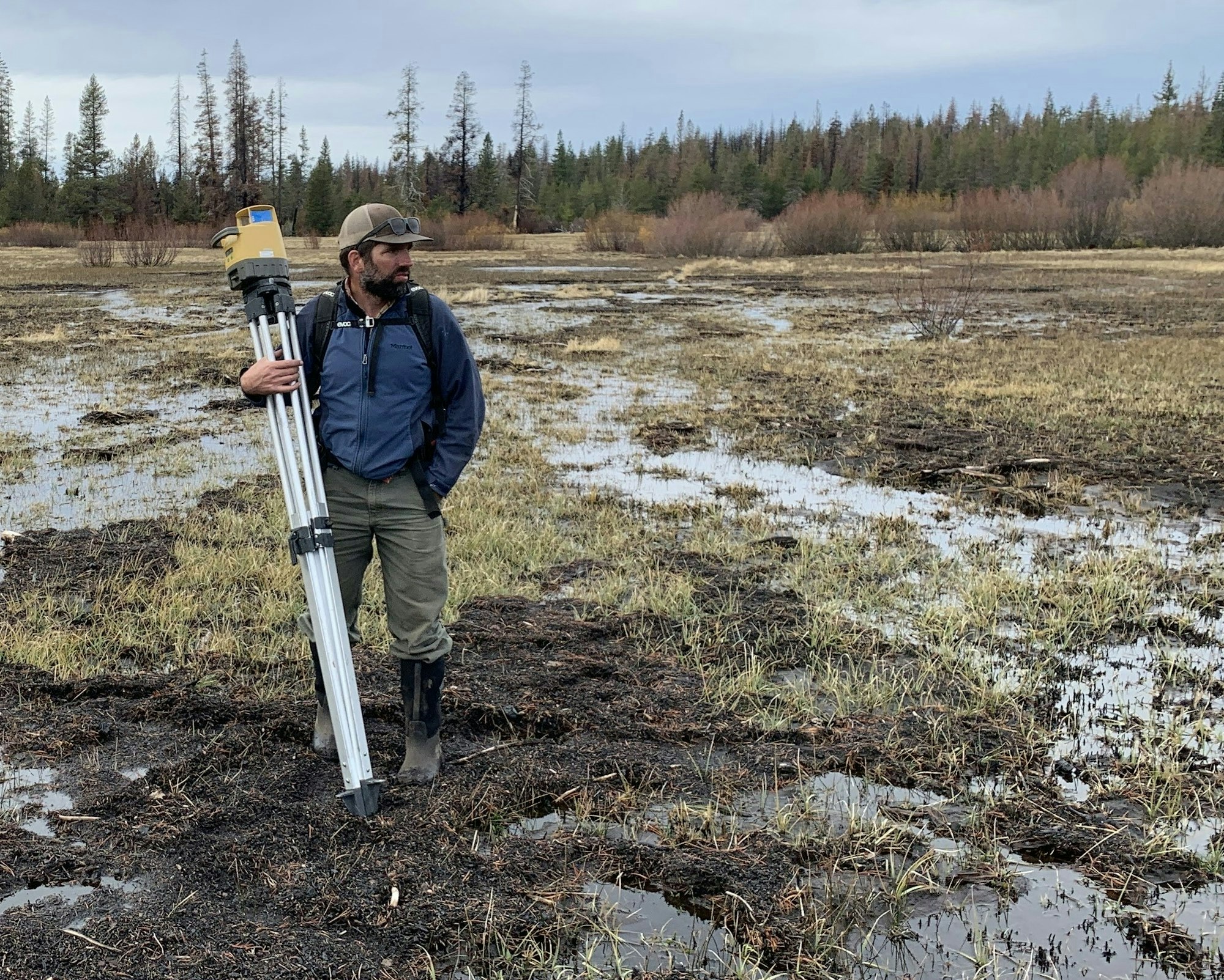 A person standing in a wet, grassy field, holding a tripod with equipment. Trees are visible in the background.