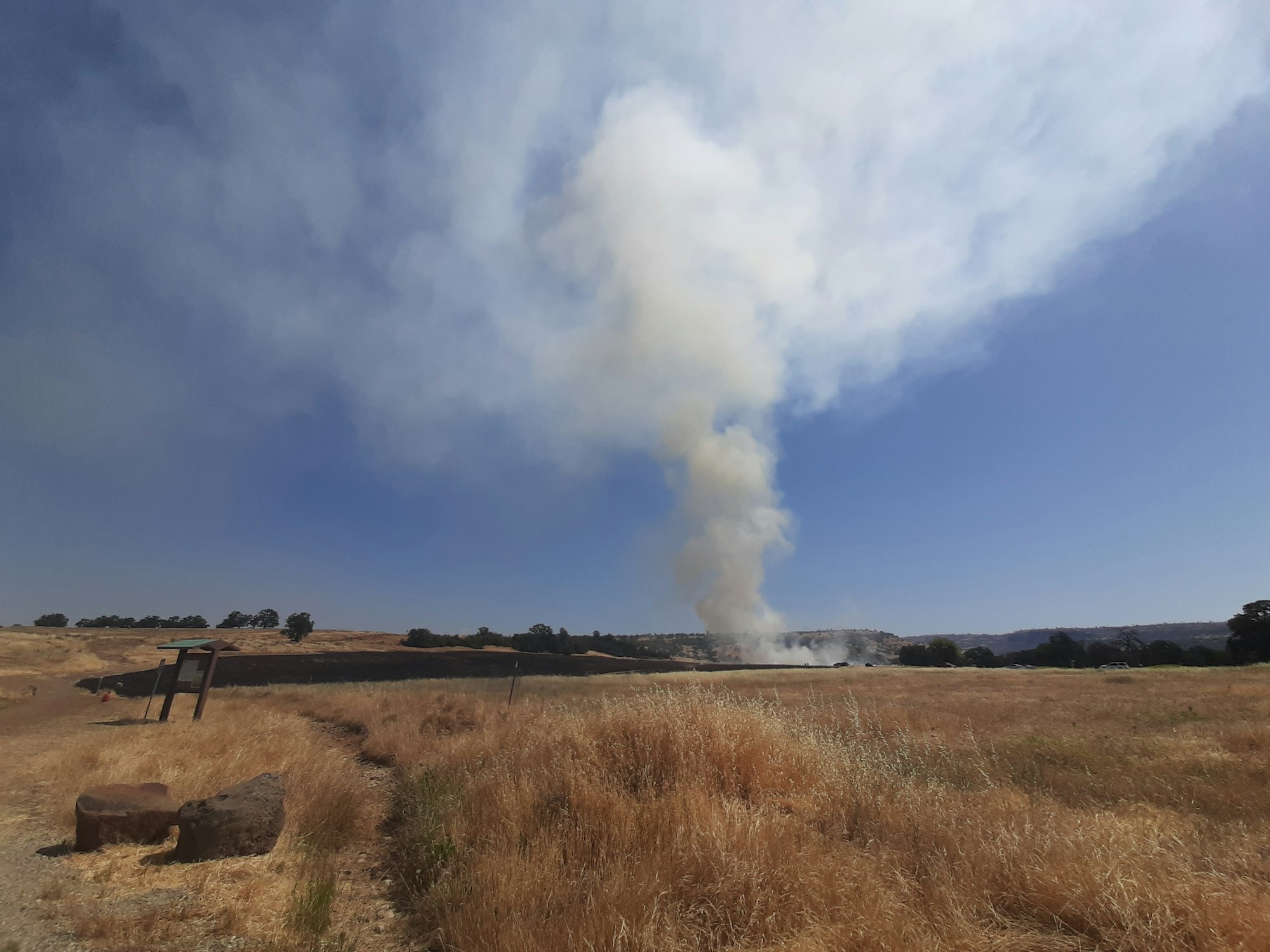 The image shows a plume of smoke rising into a clear blue sky over a dry, grassy landscape.