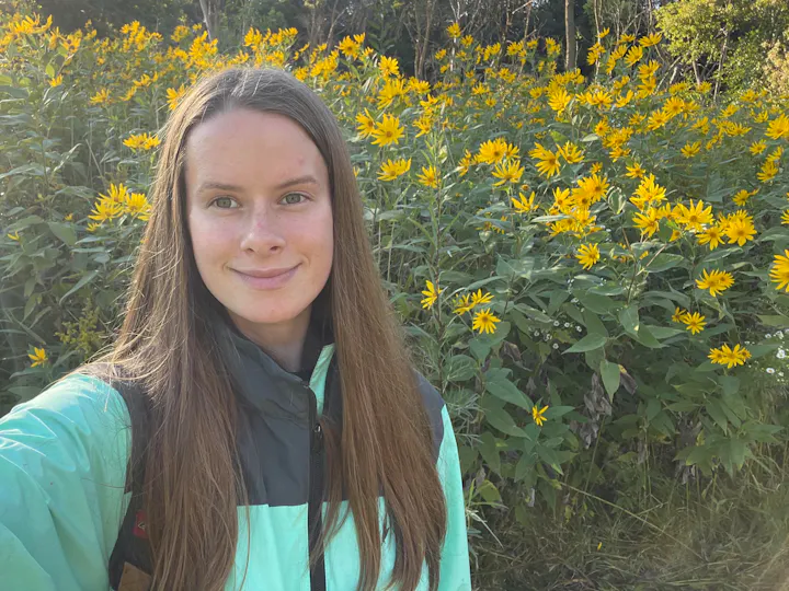 A person with long hair smiles in a field of bright yellow flowers while wearing a green and gray jacket.