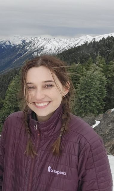 A person in a purple jacket smiling in a snowy mountain landscape with trees.