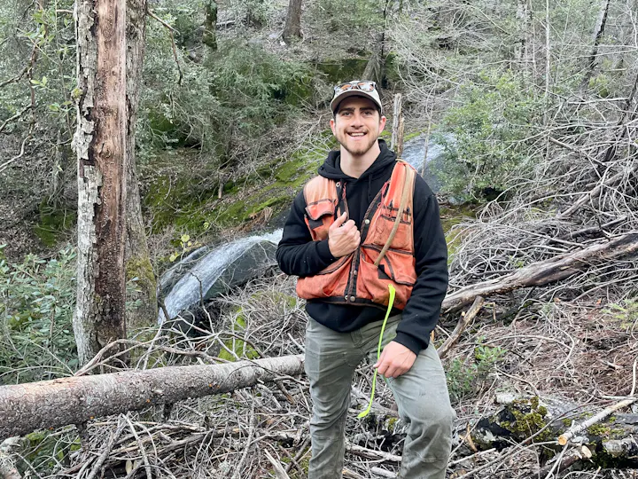 A person in an orange vest stands in a forested area, smiling, with a measuring tool in hand and trees in the background.