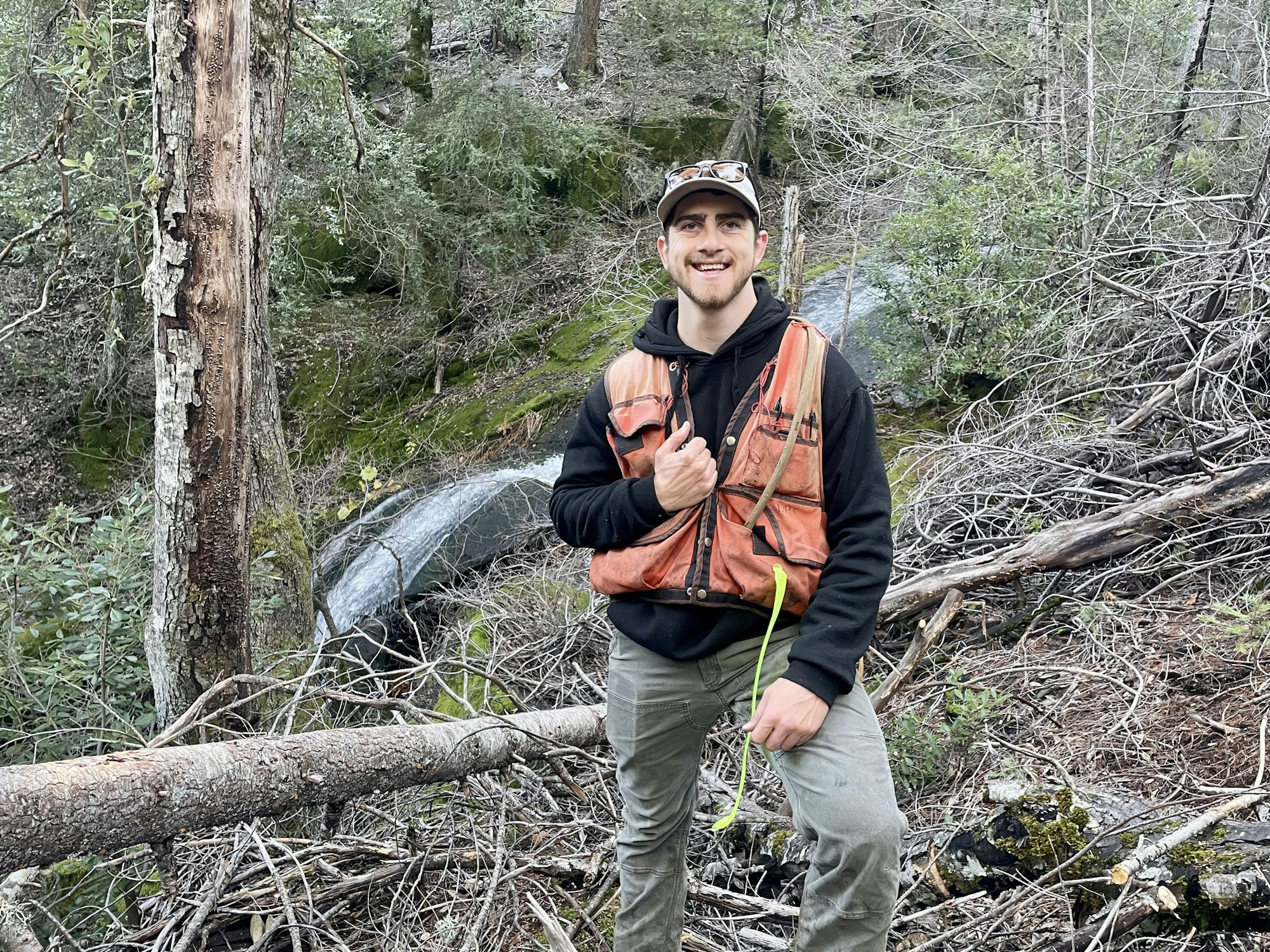 A person in an orange vest stands in a forested area, smiling, with a measuring tool in hand and trees in the background.