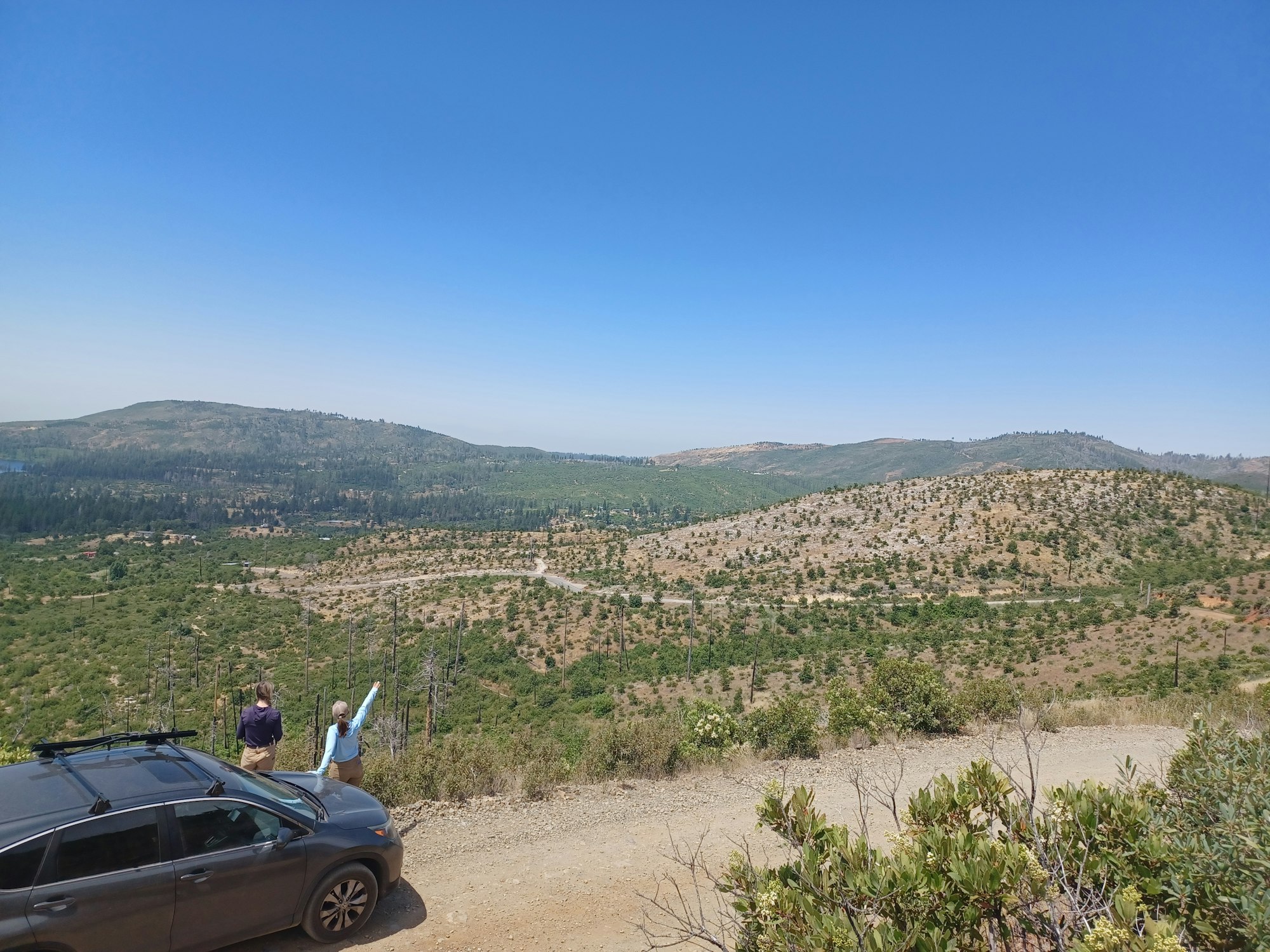 A car parked on a dirt road with two people looking at a hilly landscape under a clear blue sky.