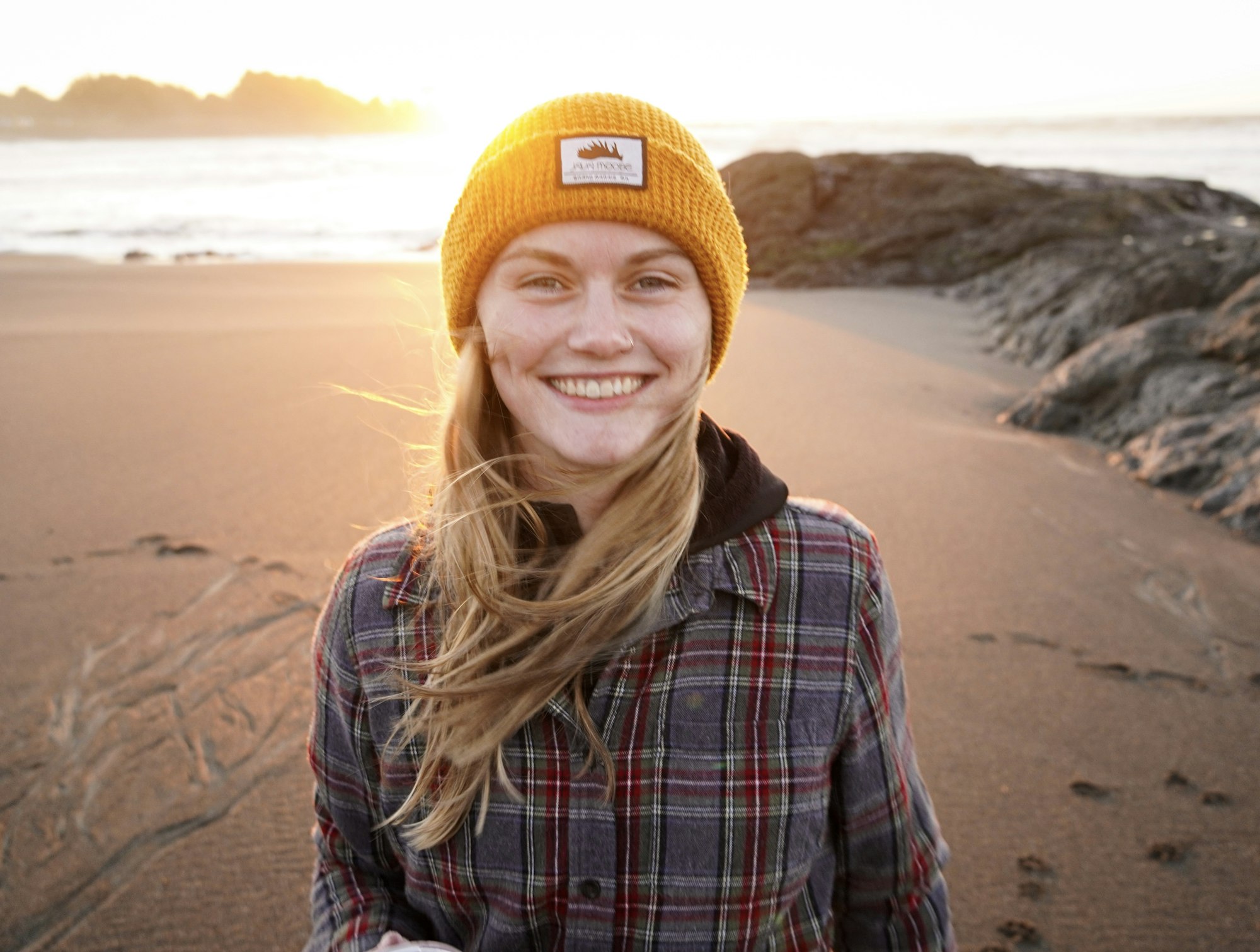 A person smiling on a beach, wearing a yellow beanie and plaid shirt, with the sun setting in the background.