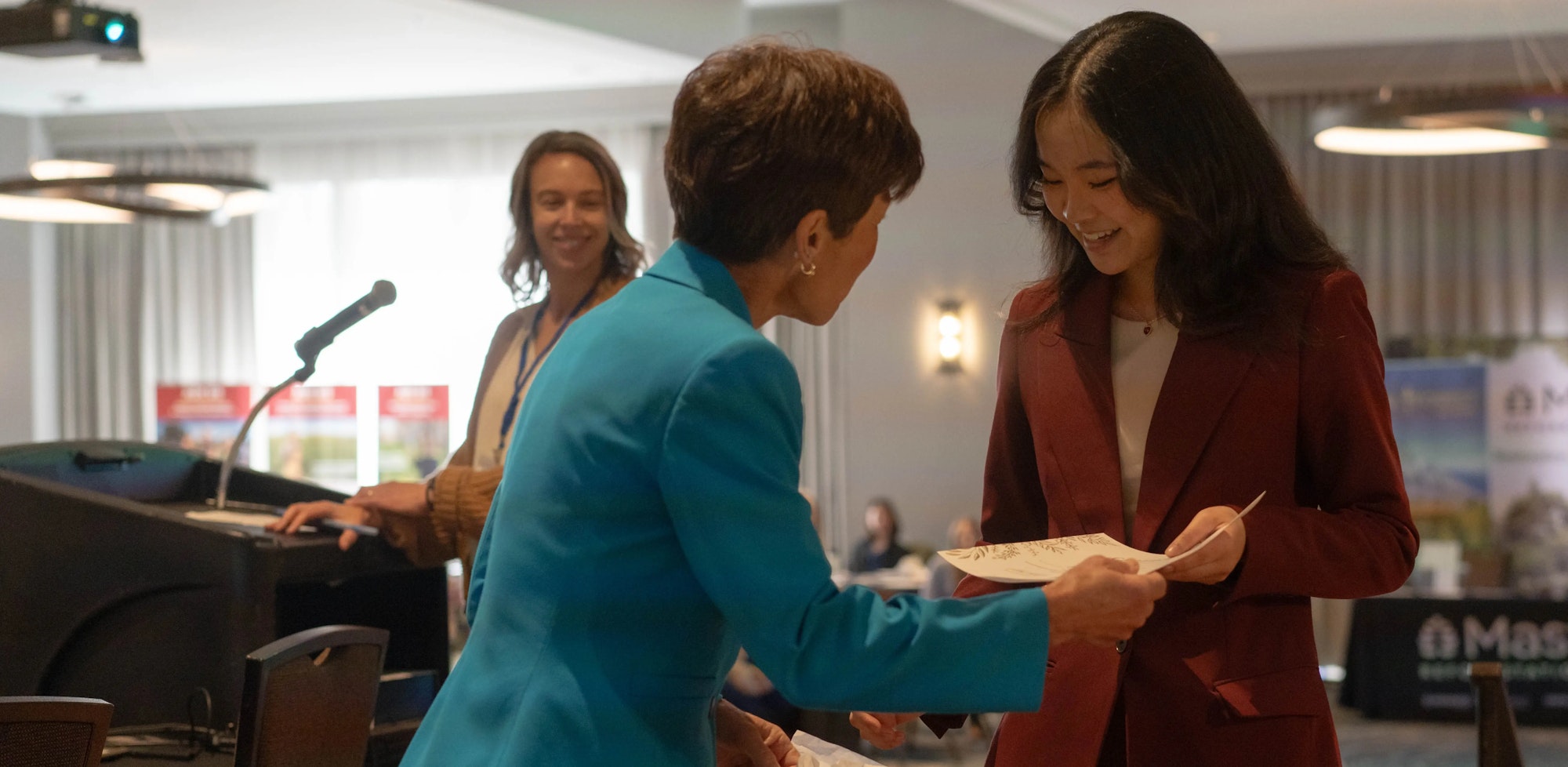 Three people at an event, with one woman in blue handing a certificate to a young woman in red, and another person smiling by a podium.