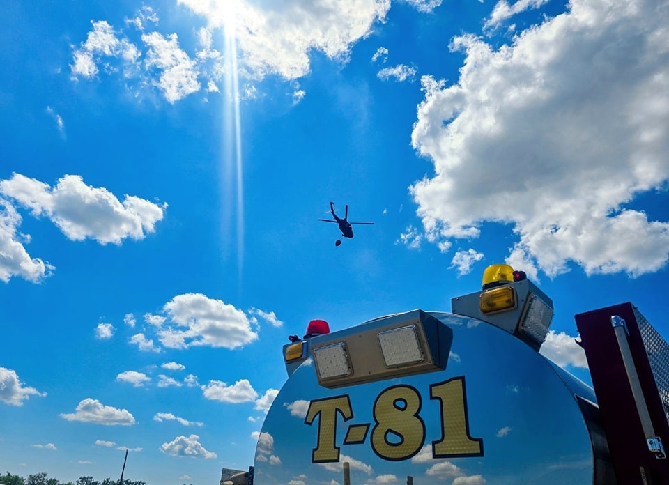 Helicopter in flight, blue sky with clouds, part of a fire truck labeled "T.81" visible in the foreground.