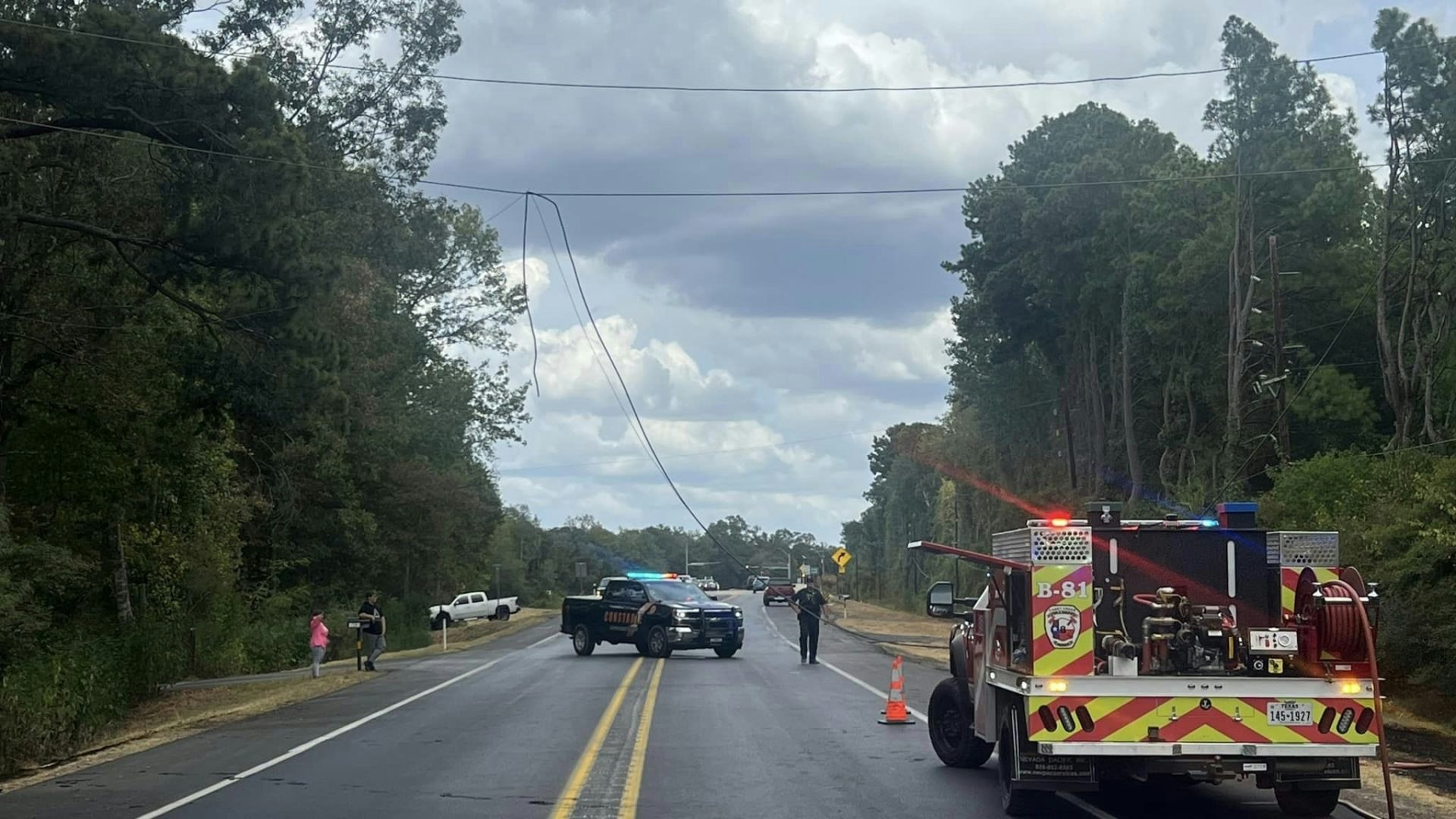 Emergency responders manage a scene on a road, with a fire truck and police blocking traffic near downed power lines.