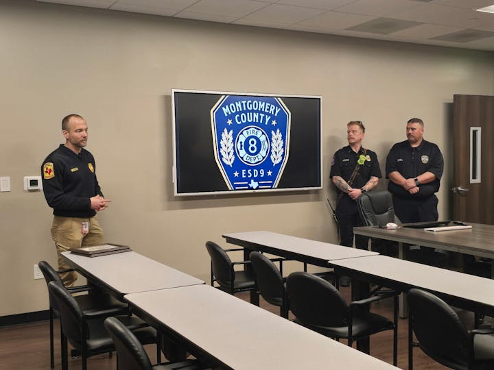 A presentation with three men in uniform, a screen displaying "Montgomery County Fire Dept. ESD 9," and empty desks in a classroom.