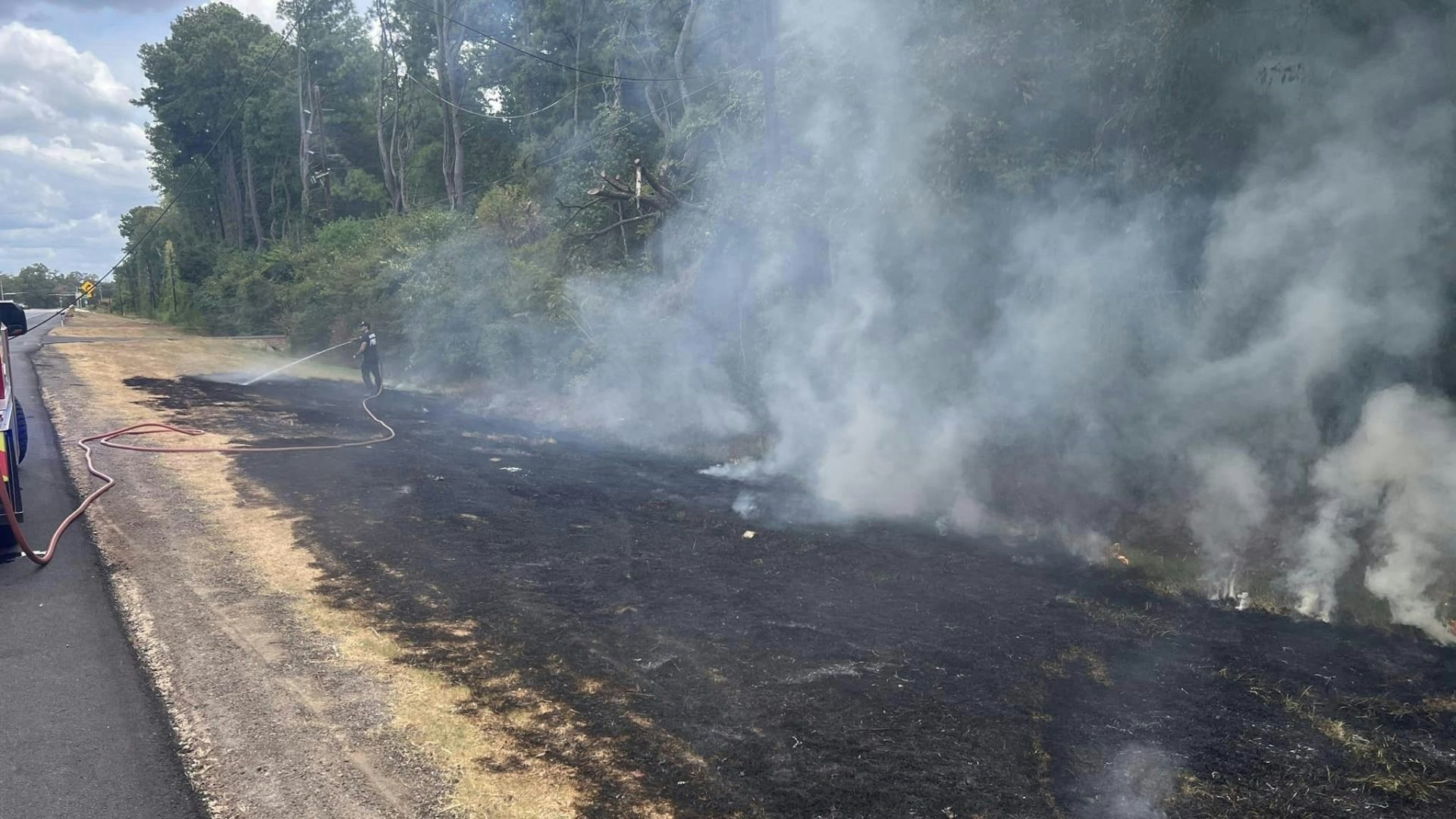 The image shows a person extinguishing smoke from a burnt area along a road, with a smoky background and fire tools visible.