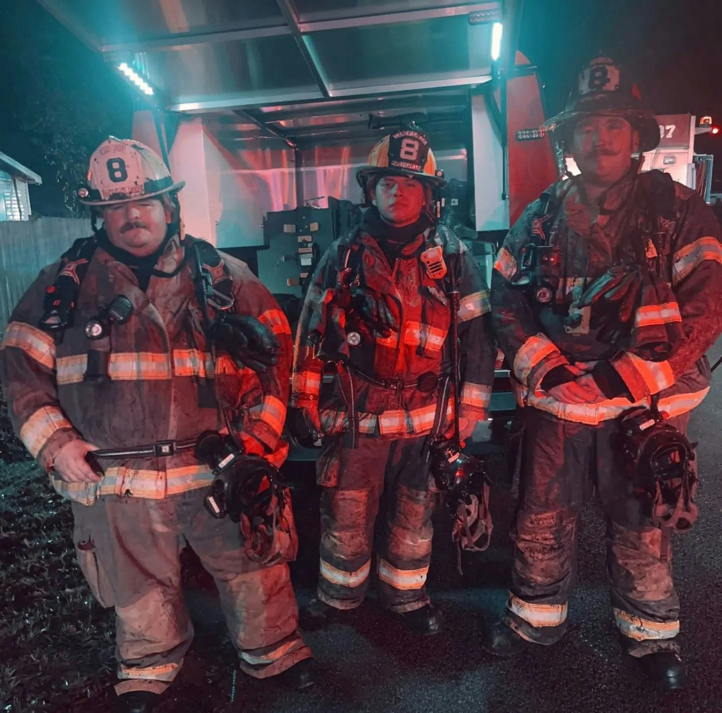 Three firefighters in gear, standing together in a dimly lit setting with red and blue lighting, showcasing teamwork.