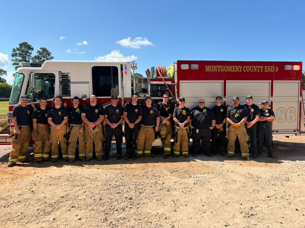 A group of firefighters stands together in front of a fire truck, all in gear, under a clear blue sky.