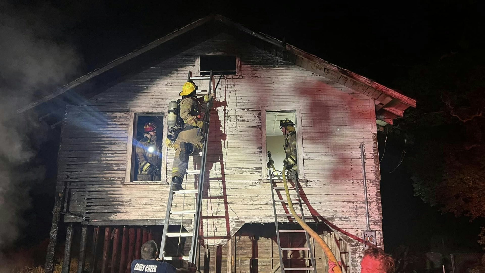 Firefighters are responding to a blaze in an old house at night, using ladders and hoses to combat the flames.