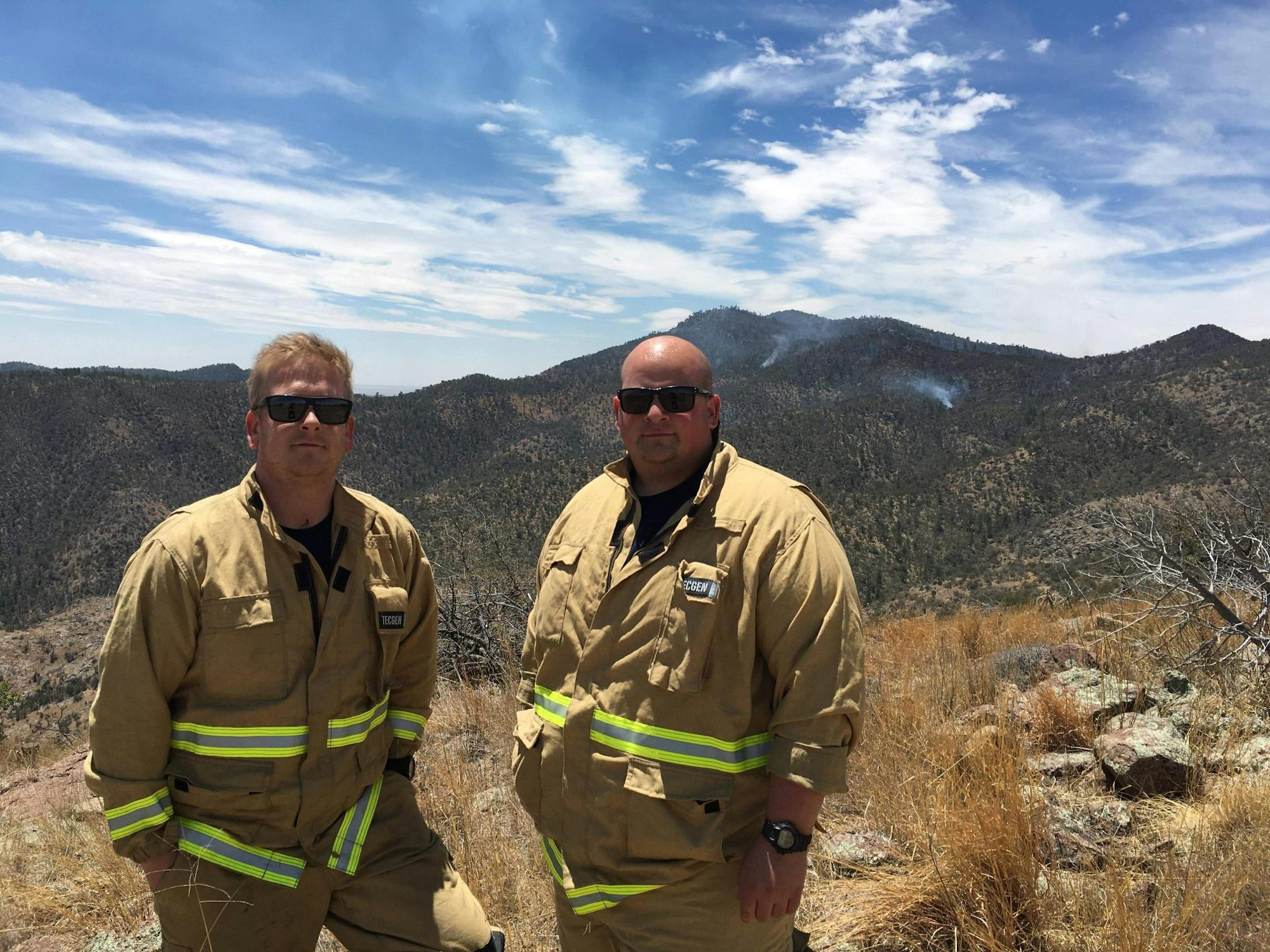 Two firefighters in tan gear with reflective stripes stand on a hillside, overlooking a smoky mountain landscape under a blue sky.