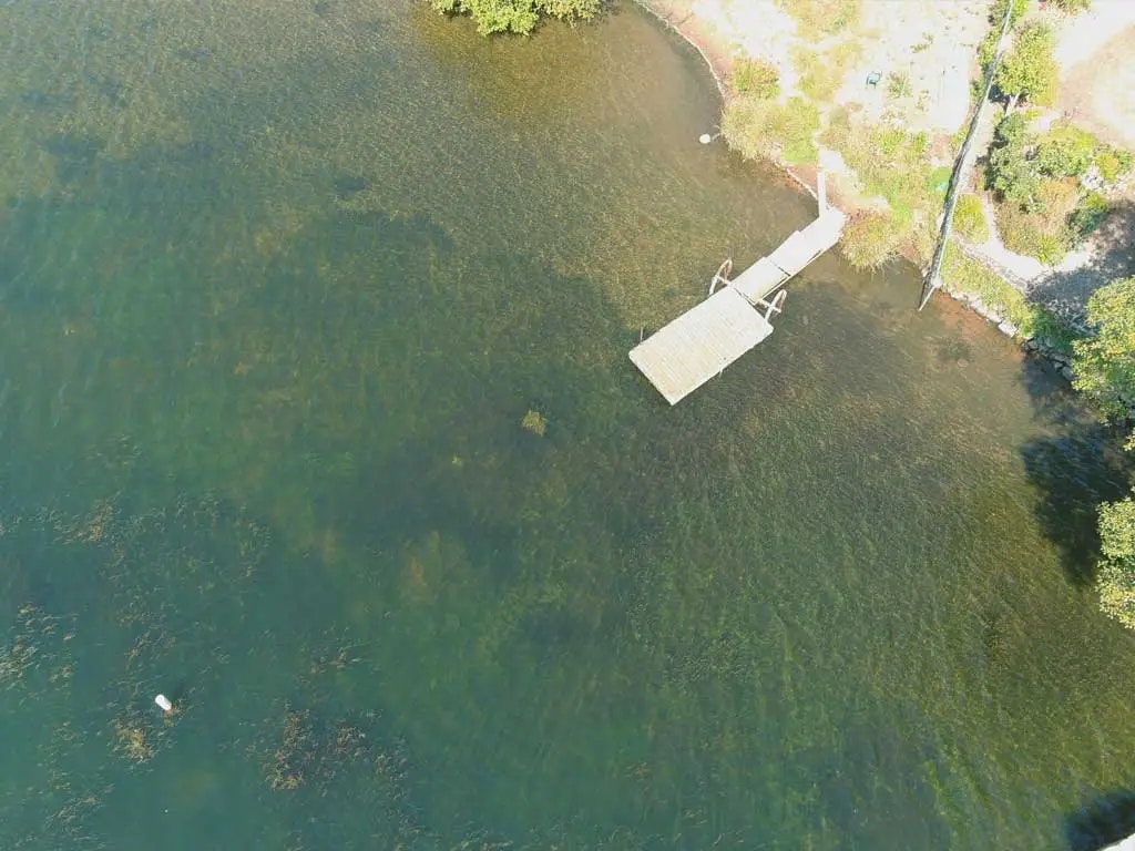 Aerial view of a dock extending into a lake with clear water, surrounded by greenery and a shoreline.