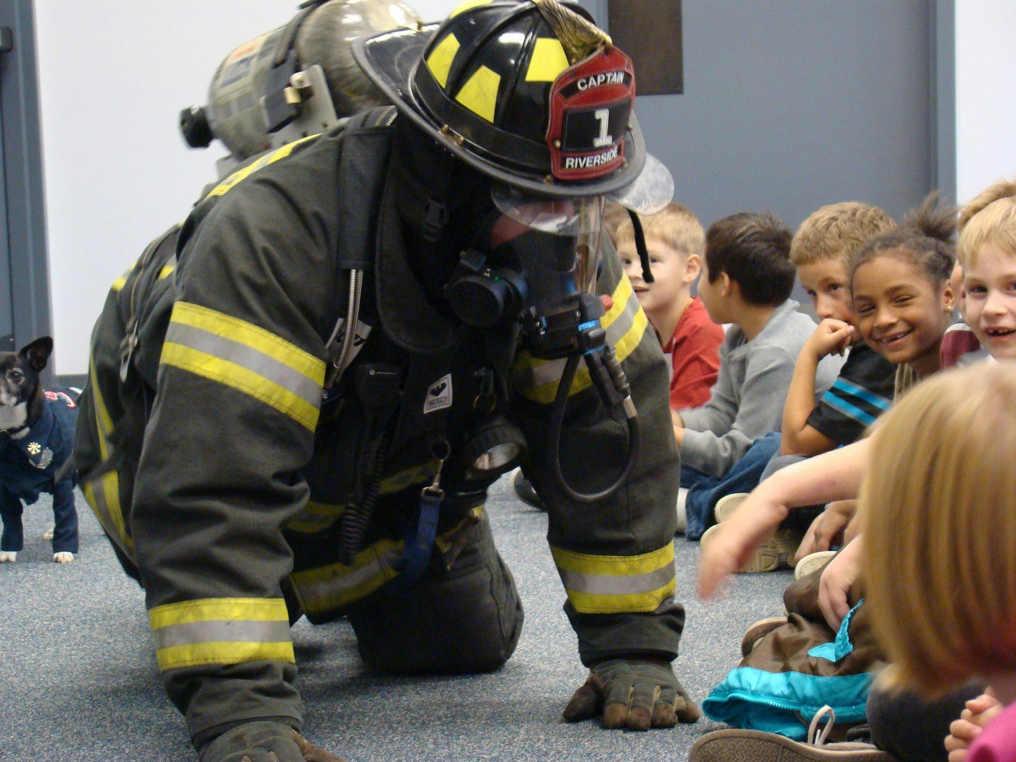 A firefighter in gear interacts with a group of smiling children, accompanied by a small dog in a sweater.
