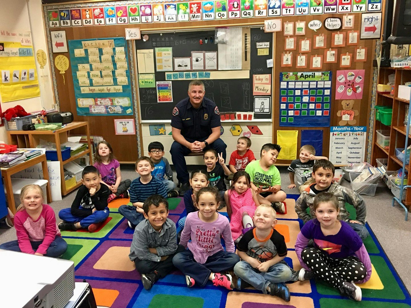 A smiling group of children sits on a colorful carpet in a classroom with a uniformed adult, likely a guest or teacher.