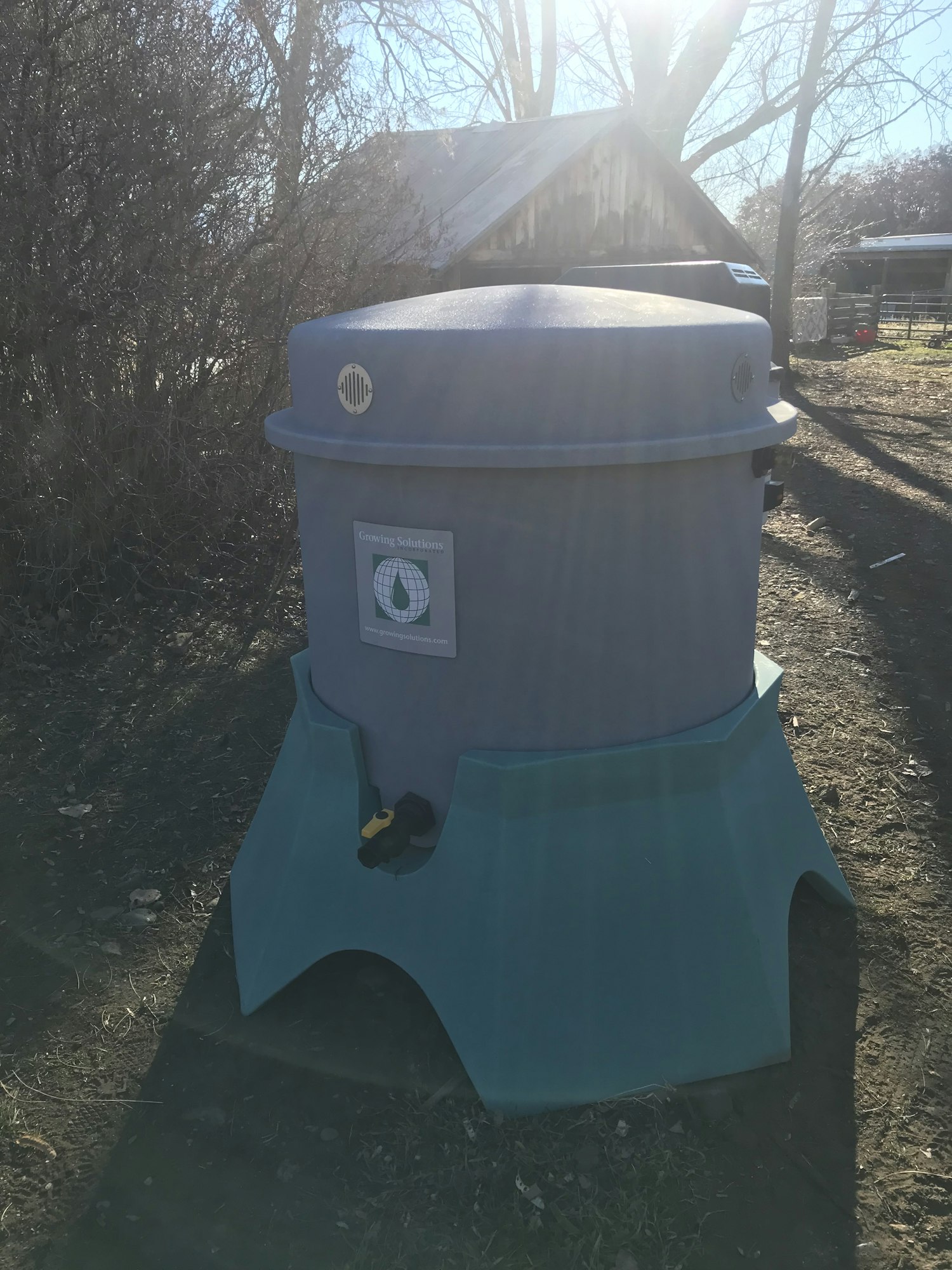 A sideways photo of a blue rain barrel with a spigot, placed on a stand outside with trees in the background.