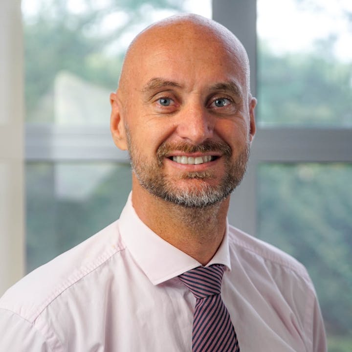 A smiling man with a bald head, light pink shirt, and striped tie poses for the camera against a blurred background.