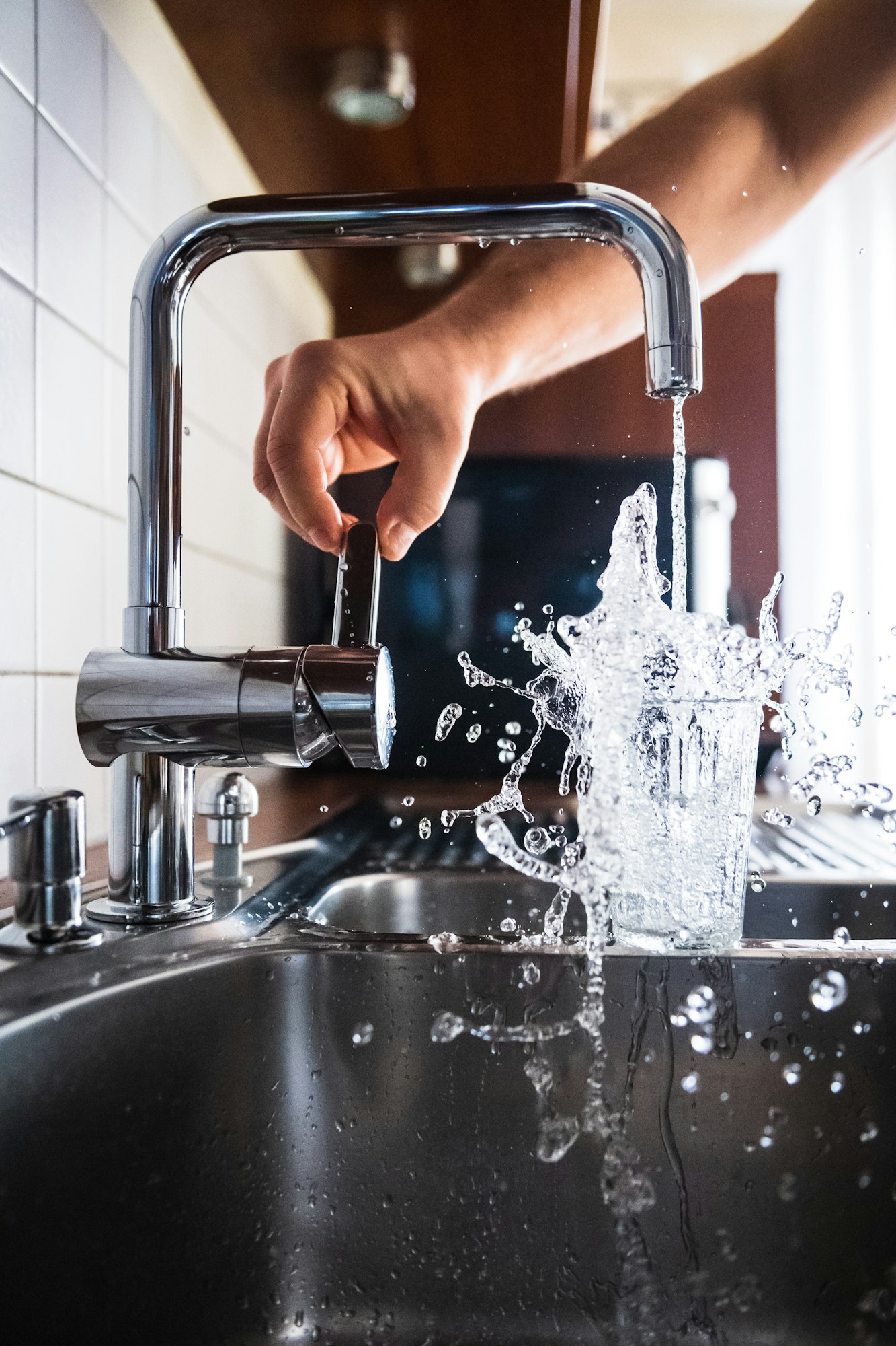 A hand operates a faucet, pouring water into a glass, creating splashes in a kitchen sink.