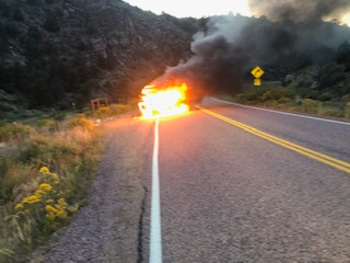 A vehicle engulfed in flames on a mountain road with smoke rising and warning road signs visible.