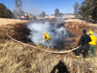 Firefighters extinguishing a smoldering field with a hose, smoke rising, fire truck in the background, and a clear blue sky.