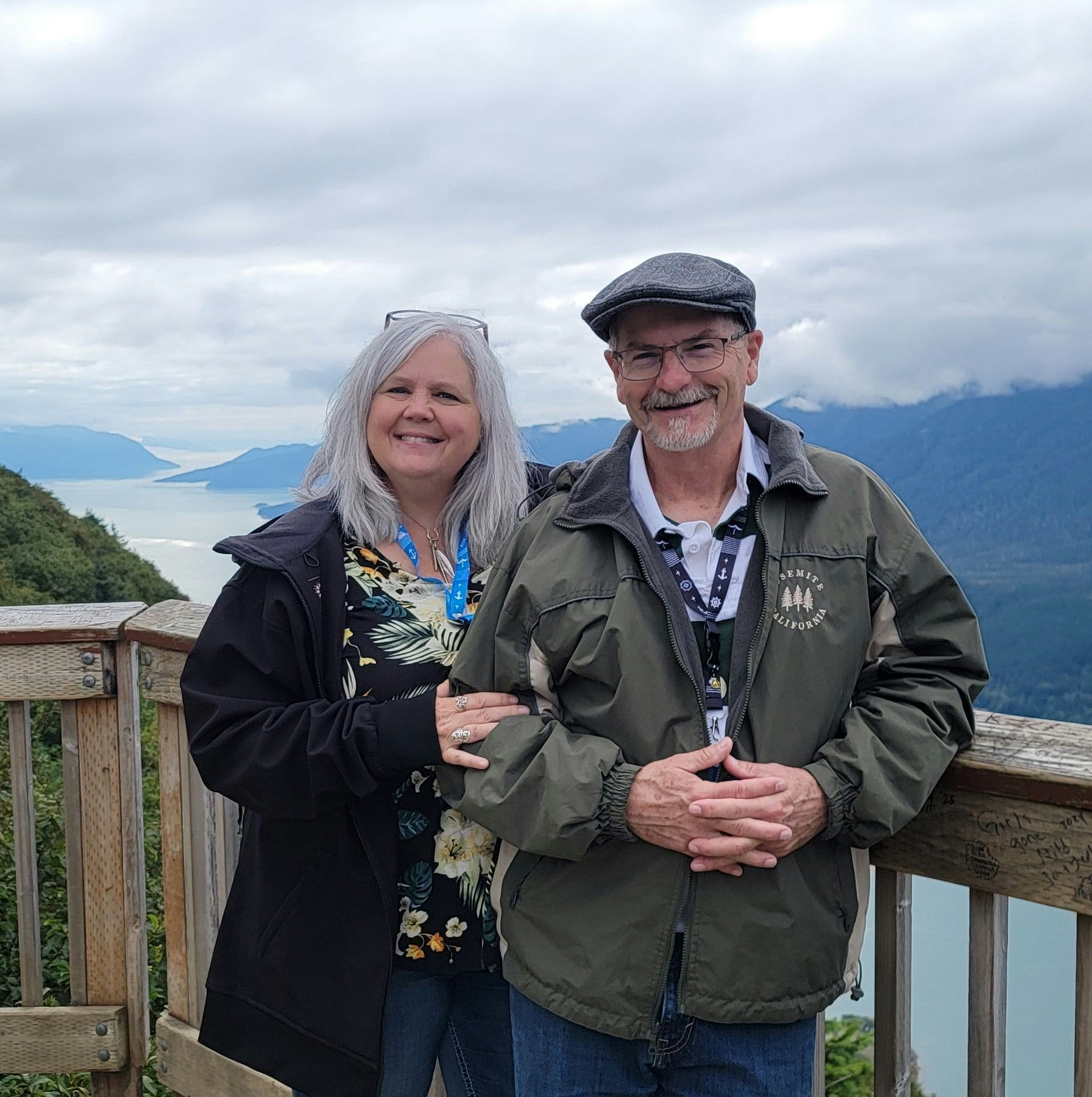 A couple stands on a wooden deck with a scenic, mountainous background under a cloudy sky.